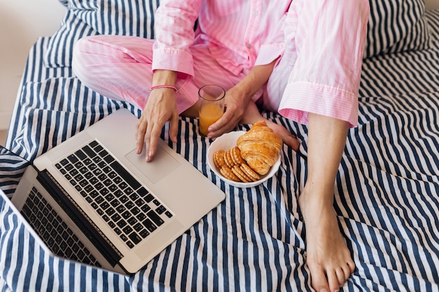 Vue de haut sur la jeune jolie femme blonde en pyjama rose assis sur le lit avec ordinateur portable prenant le petit déjeuner