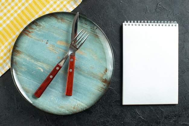 Vue horizontale des couverts de repas en croix sur une assiette bleue et une serviette jaune à côté d'un ordinateur portable sur une surface sombre