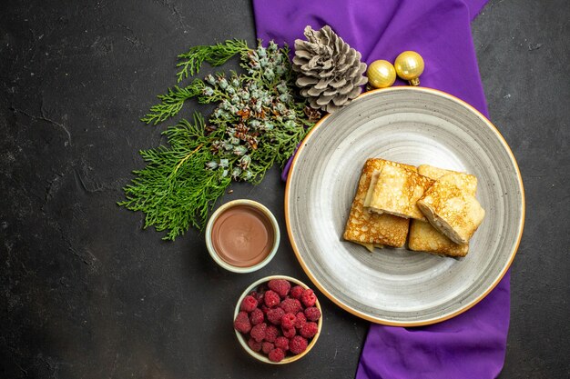 Vue horizontale de délicieuses crêpes sur une plaque blanche accessoires de décoration chocolat et framboise sur serviette violette sur fond noir