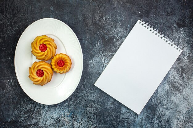 Vue horizontale de délicieux biscuits sur une assiette blanche et un cahier sur une surface sombre
