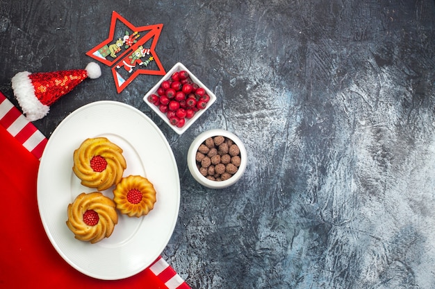 Vue horizontale de délicieux biscuits sur une plaque blanche sur une serviette rouge et un chapeau de père Noël et des chocolats dans des pots blancs sur une surface sombre