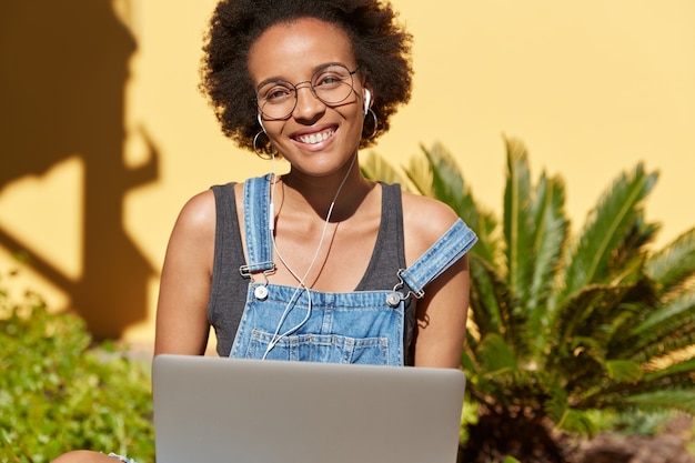 Photo gratuite vue horizontale du jeune noir avec un sourire à pleines dents, aime les blogs, travaille sur un ordinateur portable, écoute leçon audio avec des écouteurs modernes, modèles à l'extérieur, porte des lunettes optiques pour la correction visuelle