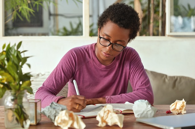 Vue horizontale du processus de travail des plans de femme à la peau sombre, notetats informations dans le bloc-notes, écrit le texte, pose à l'intérieur confortable avec des papiers. Une blogueuse prend des notes pour publication