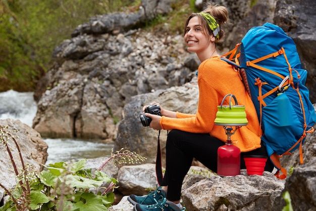 Vue horizontale de joyeuse femme heureuse se trouve près de rock pool, détient un appareil photo moderne, prépare une boisson chaude, aime le camping et les voyages, porte des vêtements de sport