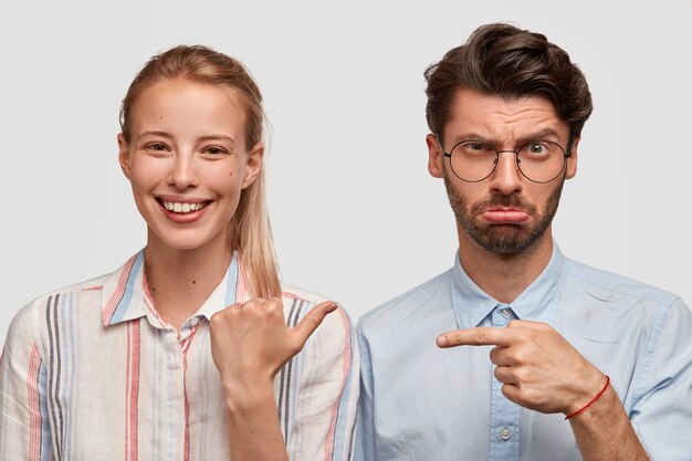 Vue horizontale de la joyeuse jeune femme européenne avec queue de cheval, habillée en chemise, pointe son mari mécontent qui a un échec, modèle contre le mur blanc. Concept de relation