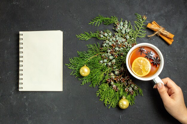 Vue horizontale de la main tenant une tasse de thé noir accessoires de Noël et citrons verts à la cannelle et cahier sur fond noir