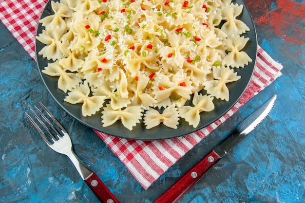 Photo gratuite vue horizontale des pâtes farfalle italiennes crues avec des légumes sur une plaque noire sur une serviette rayée rouge à côté des couverts sur le côté droit sur fond bleu