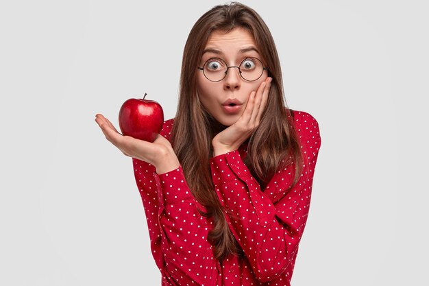 Vue intérieure d'une dame de race blanche surprise touche la joue avec étonnement, détient la pomme rouge, porte des lunettes