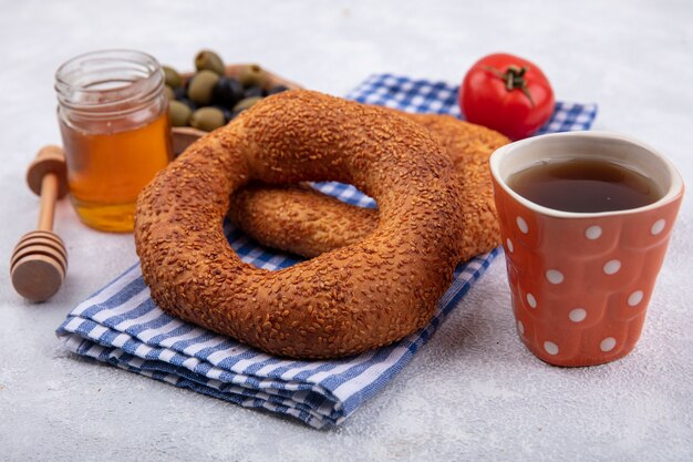 Vue latérale des bagels turcs traditionnels doux isolés sur un chiffon à carreaux avec une tomate et olives sur un bol en bois avec du miel sur un bocal en verre sur fond blanc