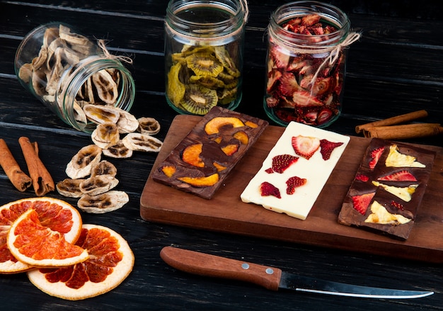 Photo gratuite vue latérale des barres de chocolat noir et blanc sur une planche à découper en bois avec divers fruits en tranches séchées sur fond noir