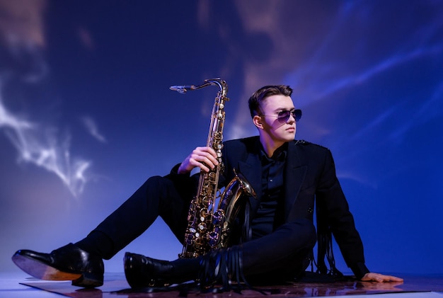 Photo gratuite vue latérale d'un beau musicien masculin portant un costume noir à la mode et des lunettes de soleil assis sur le sol et tenant un saxophone doré tout en détournant sérieusement les yeux et en posant