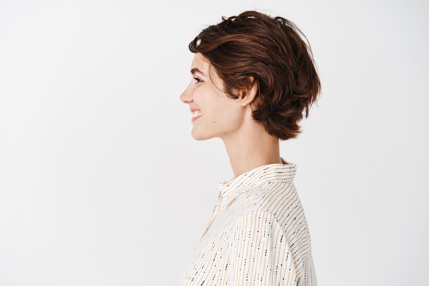 Vue latérale d'une belle jeune femme avec une peau propre et saine, aspect naturel sans maquillage, regardant à gauche et souriante, debout sur un mur blanc