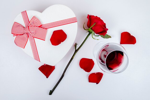 Vue latérale d'une boîte-cadeau en forme de coeur et d'un verre de vin de couleur rouge rose et pétales sur fond blanc