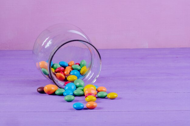 Vue latérale de bonbons colorés dispersés dans un bocal en verre sur fond violet avec copie espace