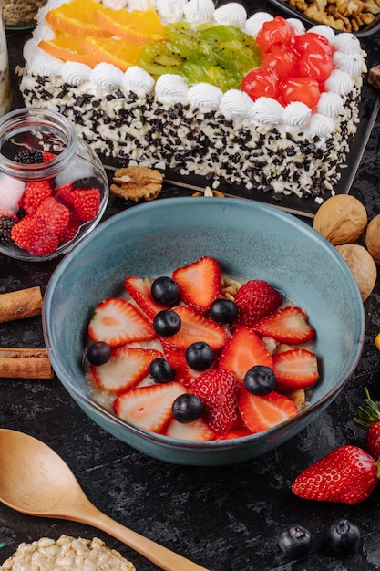 Vue latérale de la bouillie d'avoine avec des fraises et des bleuets en tranches dans un bol en céramique et un gâteau aux fruits avec de la crème fouettée sur la table