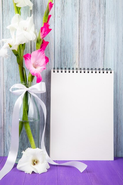 Vue latérale d'un carnet de croquis avec des fleurs de glaïeul de couleur blanche et rose dans une bouteille en verre sur fond de bois gris