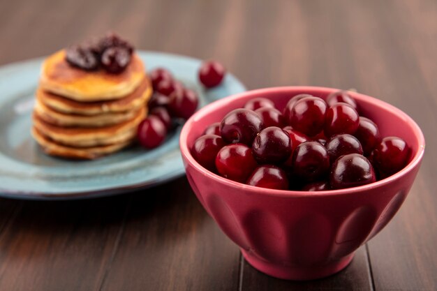 Vue latérale des cerises dans un bol et assiette de crêpes et cerises sur fond de bois