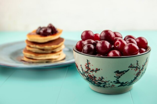 Vue latérale des cerises dans un bol avec des crêpes en plaque sur fond bleu