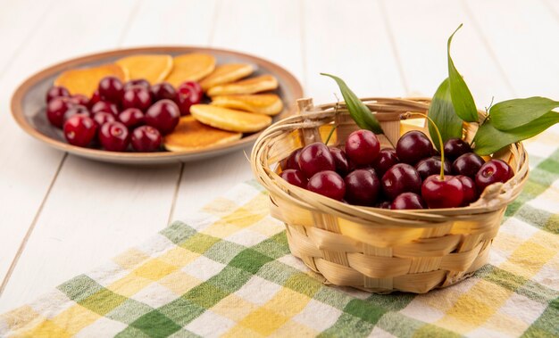 Vue latérale des cerises dans le panier sur tissu à carreaux et assiette de crêpes et cerises sur fond de bois