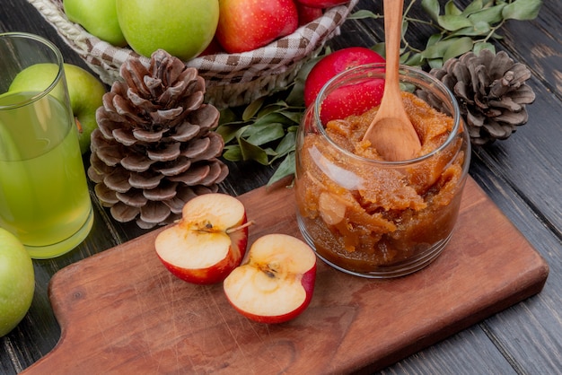 Vue latérale de la confiture de pommes dans un bocal en verre avec une cuillère en bois et une demi-pomme coupée sur une planche à découper avec un panier de jus de pomme de pommes pomme de pin et de feuilles sur une surface en bois