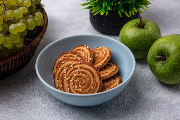 Vue latérale des cookies dans un bol avec des pommes vertes et des raisins verts sur fond blanc