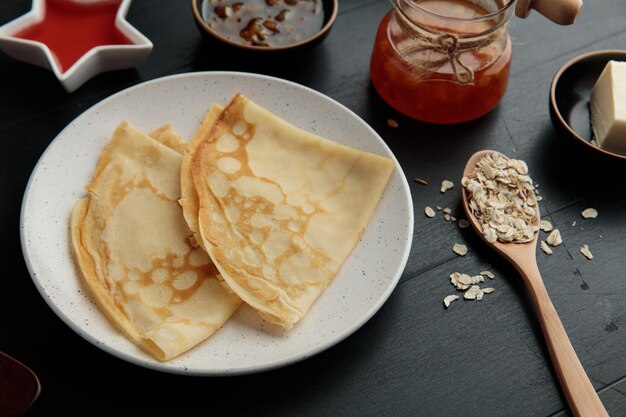 Vue latérale des crêpes dans une assiette avec de la confiture dans un bocal et dans des bols avec une cuillerée de beurre d'avoine sur fond noir