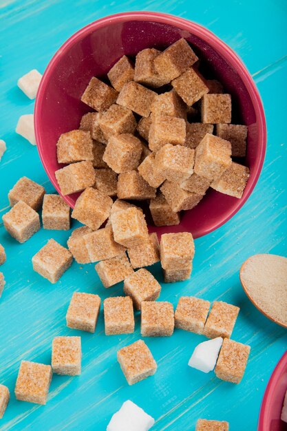 Vue latérale des cubes de sucre brun éparpillés dans un bol rose sur fond de bois bleu