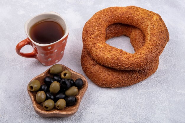 Vue latérale de délicieux bagels turcs au sésame avec une tasse de thé et d'olives sur un bol en bois sur fond blanc