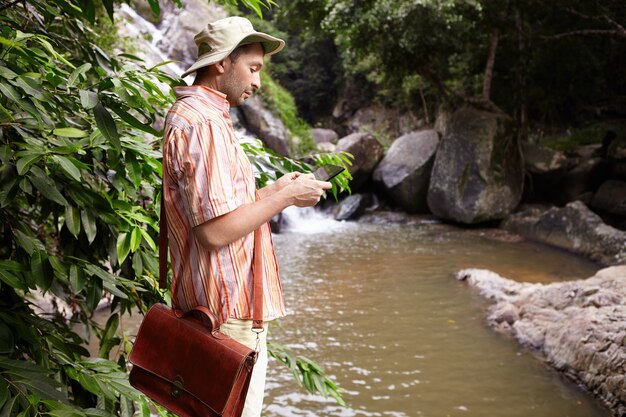 Vue latérale du biologiste masculin avec sac en cuir au travail sur le terrain, effectuer l'analyse de l'eau debout à la rivière de montagne et prendre des photos à l'aide de tablette numérique avec cascade et rochers