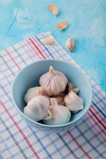 Vue latérale du bol de bulbes d'ail avec des gousses d'ail sur un tissu écossais sur fond bleu