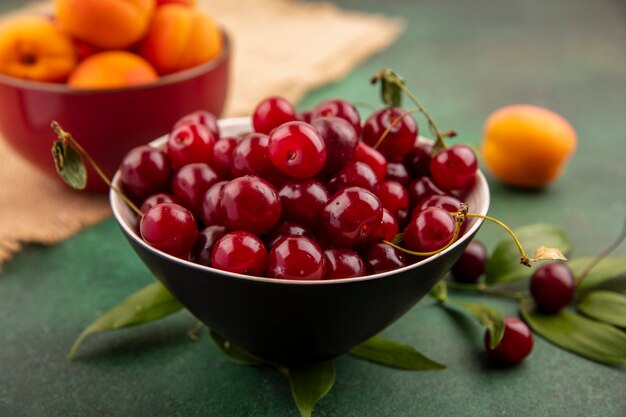 Vue latérale du bol de cerises avec bol d'abricots sur un sac et de feuilles sur fond vert