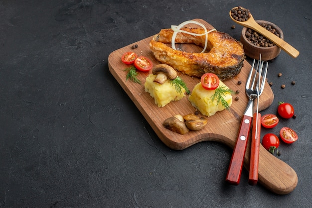 Vue latérale du délicieux poisson frit et champignons tomates verts sur une planche à découper en bois couverts mis poivre sur surface noire