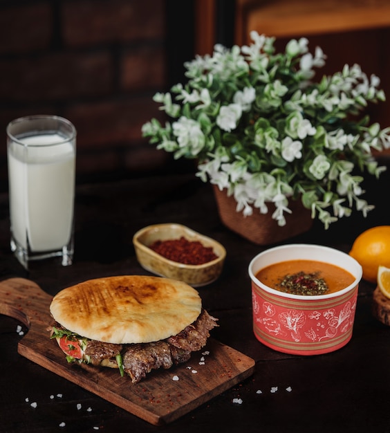 Vue latérale du doner kebab dans du pain pita sur une planche de bois servi avec une soupe au linteau et une boisson ayran sur le tableau noir