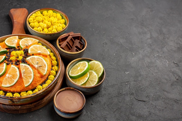 Vue latérale du gâteau et des bols de citron vert de bonbons au chocolat, de la crème au chocolat et des citrons verts à côté du gâteau à l'orange appétissant sur la table