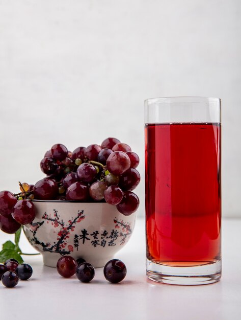 Vue latérale du jus de raisin noir dans un verre et d'un bol de raisins rouges sur fond blanc