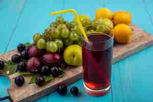 Photo gratuite vue latérale du jus de raisin noir avec tube à boire en verre et nectacots pluots raisin sur planche à découper sur fond bleu