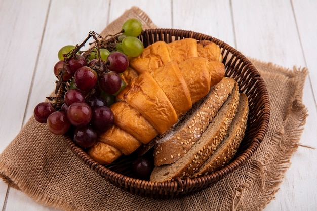Photo gratuite vue latérale du pain comme croissant et tranches de pain d'épis brun épépiné avec du raisin dans le panier sur un sac sur fond de bois