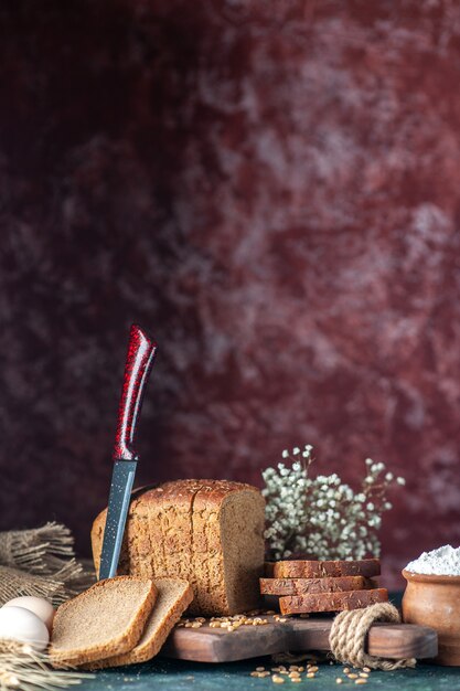 Vue latérale du pain noir diététique blés sur planche à découper en bois couteau fleur oeufs farine dans bol serviette marron sur fond de couleurs mélangées