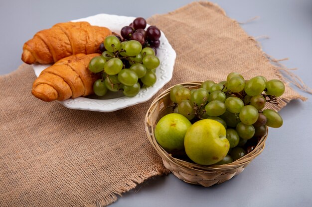 Vue latérale du panier et assiette de raisins avec pluots et croissants sur un sac sur fond gris