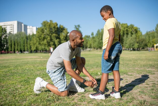 Vue latérale du père africain et de son fils debout sur le terrain. Homme souriant debout sur son genou sur l'herbe attachant des lacets sur des baskets pour enfants, les deux se regardant. Soins aux parents et concept d'activité sportive