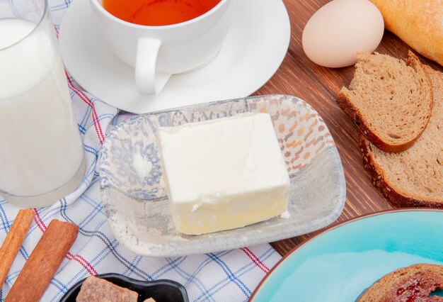 Vue latérale du petit-déjeuner avec des tranches de pain de seigle au beurre maculées de confiture dans une assiette de thé au lait à la cannelle sur un tissu à carreaux et une table en bois