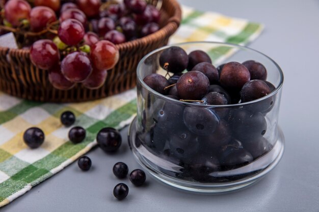 Vue latérale du raisin noir dans le panier sur tissu à carreaux et baies de raisin dans un bol sur fond gris