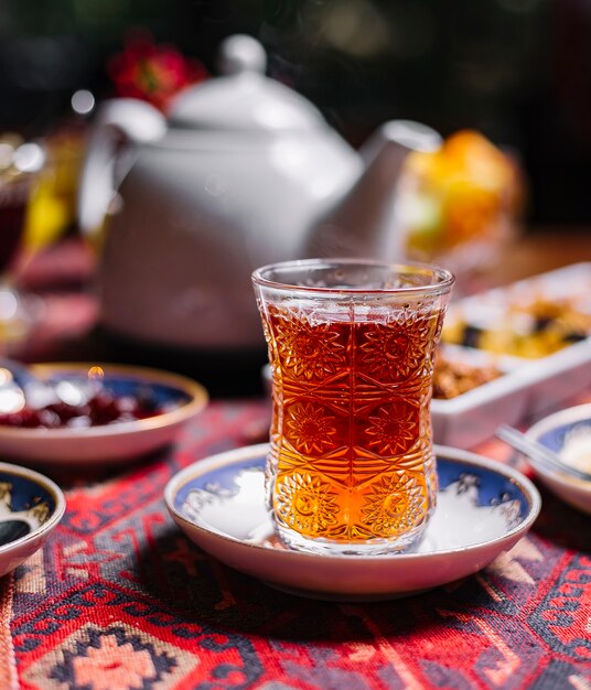 Vue latérale du thé noir dans un verre en forme de poire sur une soucoupe avec des bonbons et une théière sur la table