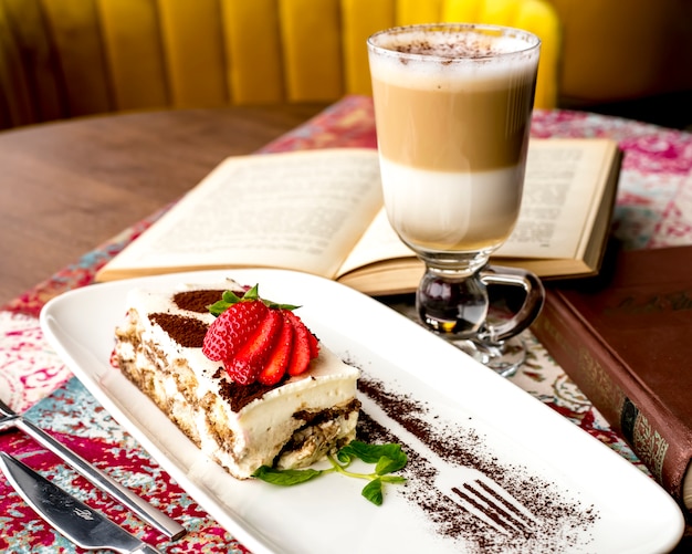 Photo gratuite vue latérale du tiramisu décoré de fraises tranchées et de poudre de cacao sur une assiette servie avec un verre de latte macchiato sur la table