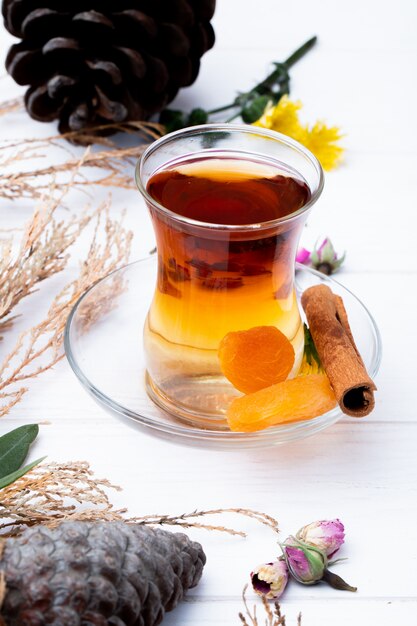 Vue latérale du verre de thé armudu aux abricots secs, bâton de cannelle et boutons de rose secs éparpillés sur blanc