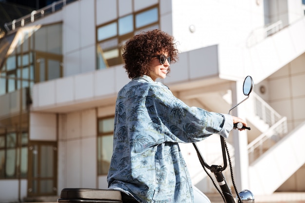 Vue latérale de l'élégante femme frisée en lunettes de soleil posant sur une moto moderne à l'extérieur