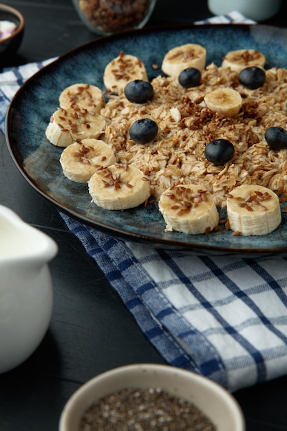 Photo gratuite vue latérale de la farine d'avoine aux noix de prunellier de banane dans une assiette sur un tissu à carreaux avec du lait et des graines de chia sur fond noir