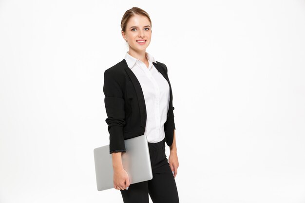 Vue latérale d'une femme d'affaires blonde souriante posant en studio avec un ordinateur portable à la main et sur un mur blanc