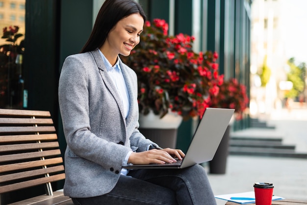 Photo gratuite vue latérale d'une femme d'affaires utilisant un ordinateur portable à l'extérieur tout en prenant un café