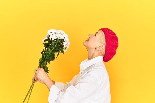 Vue latérale d'une femme âgée excitée et ravie dans un béret élégant et une chemise décontractée posant sur fond de mur de studio jaune blanc tenant des fleurs et levant les yeux comme si elle allait lancer le bouquet